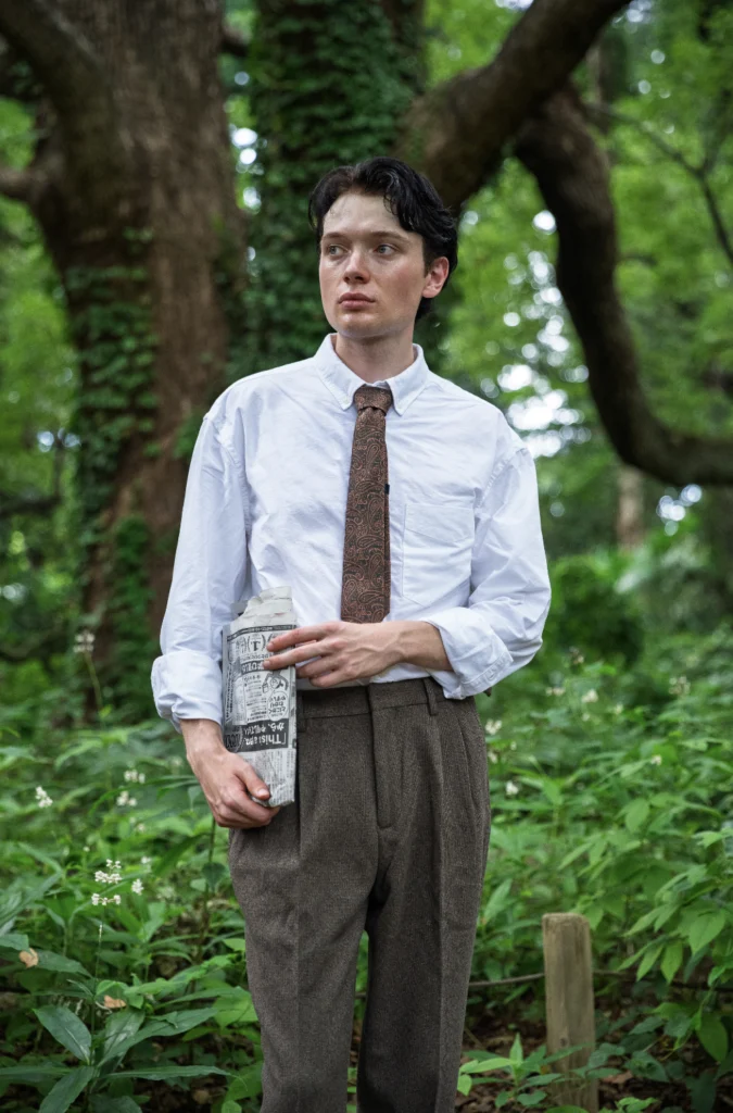 A person in a white shirt and patterned tie holding a folded newspaper among trees.