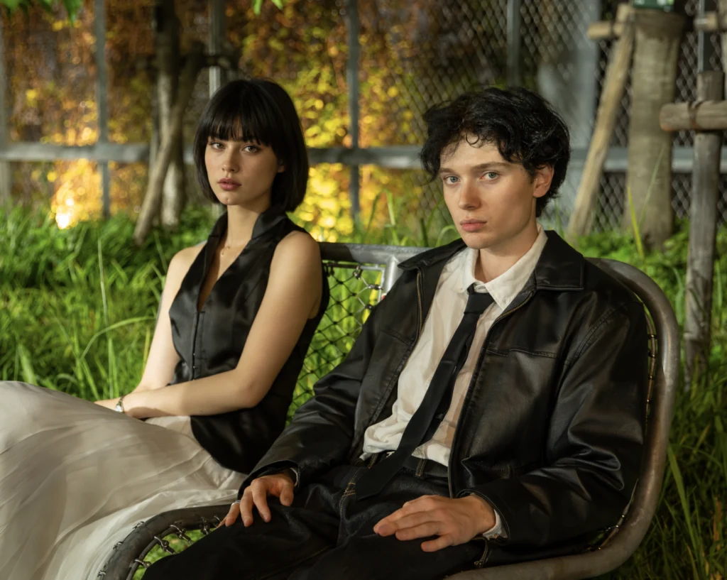 Two people in dark clothing seated beside lush greenery and a wire fence.