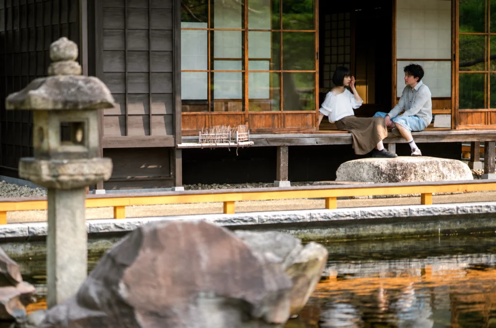 Two people seated on a wooden veranda beside a pond and traditional architecture.