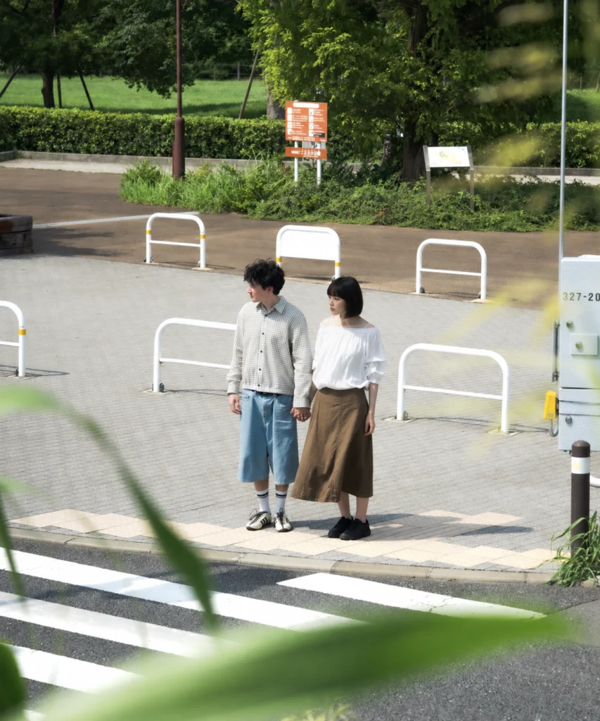 Two people holding hands beside a street crossing, framed by soft foreground foliage.